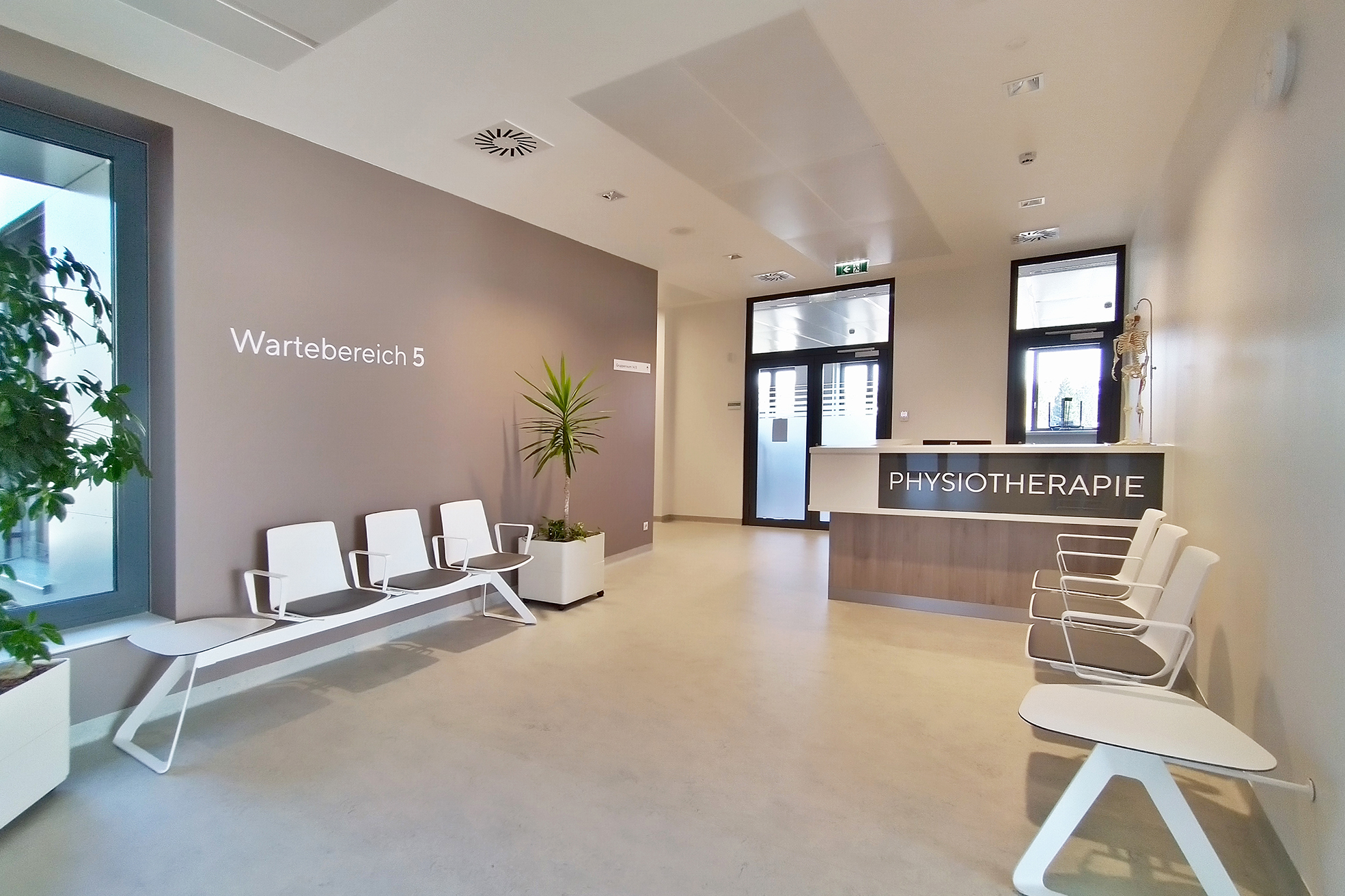 Wartezone im Therapiebereich in Braun- und Beiget&ouml;nen mit Topfpflanzen; links und rechts an der Mauer montierte Sitzreihen, im rechten hinteren Bereich das Anmeldepult mit gro&szlig;em Schriftzug Physiotherapie; Zwei Glast&uuml;ren dahinter f&uuml;hren auf einen Gang.