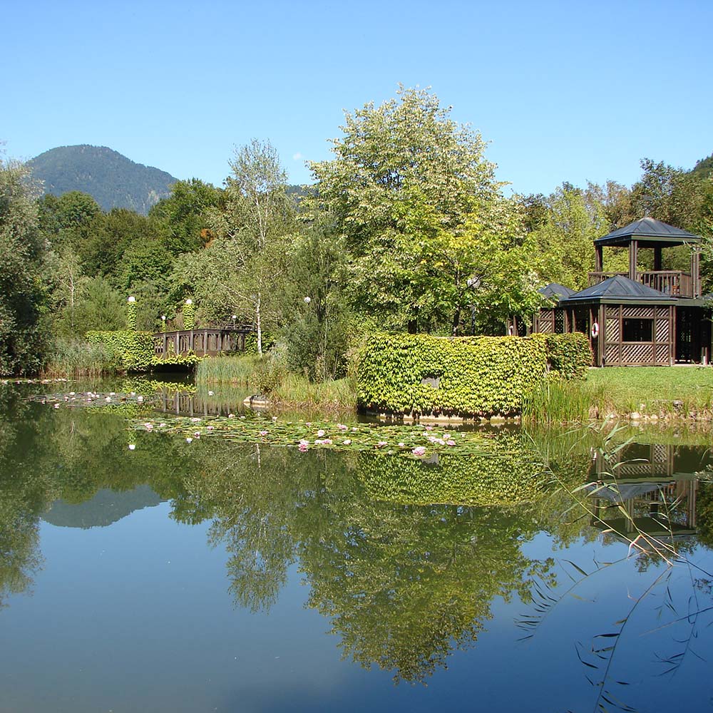 Wundersch&ouml;ne Gartenanlage des Gesundheitszentrums: im Vordergrund ein Teich mit Seerosen, am Ufer beginnt die Gartenanlage mit kleinen Holzpavillons, Wiesen, B&auml;umen und Str&auml;uchern. Eine Holzbr&uuml;cke f&uuml;hrt &uuml;ber den Deichzufluss.