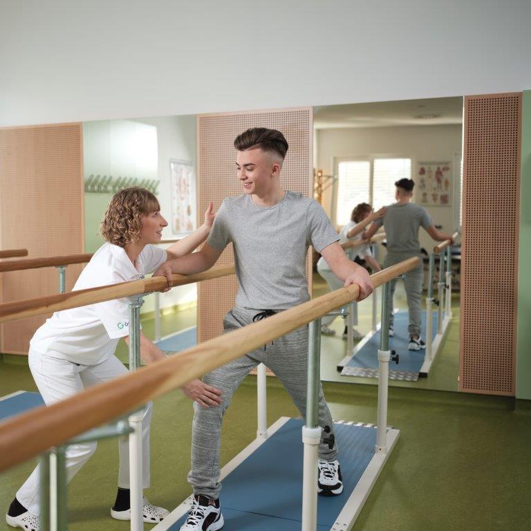 &Uuml;bung im Therapieraum: Ein Mann steht in Schrittstellung im Parallelgehbarren und h&auml;lt sich am Handlauf fest. Die Therapeutin links daneben hilft ihm, die richtige K&ouml;rperhaltung einzunehmen.