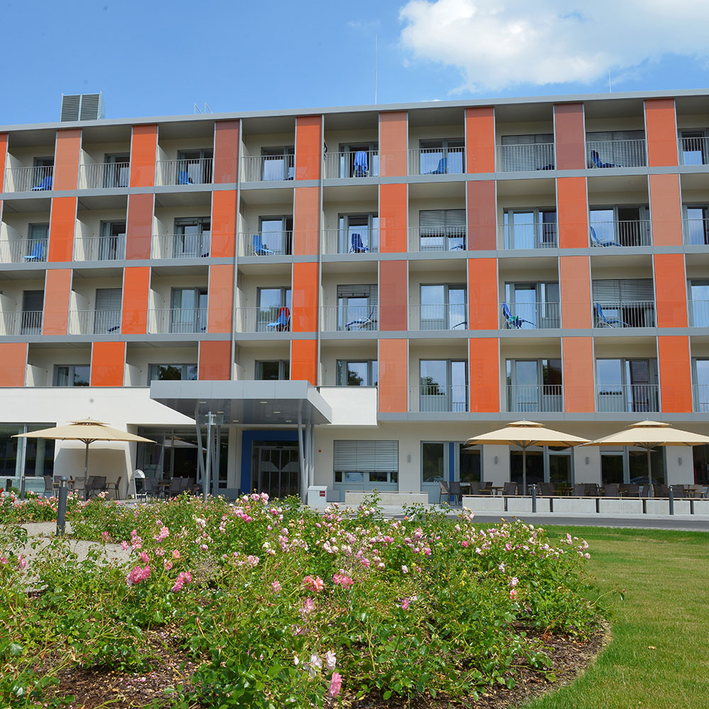 Au&szlig;enansicht Gesundheitszentrum Peterhof; Vierst&ouml;ckiger Flachdachbau mit Sicht auf Balkone mit orangen Trennw&auml;nden; links und rechts vor dem &uuml;berdachten Eingang stehen Sitzgruppen mit Sonnenschirmen; im Vordergrund wachsen Rosen und eine Wiese;
