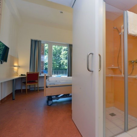 Helles Einzelzimmer mit dunklem Boden, Eckschreibtisch mit Sessel, Fernseher an der Wand gegen&uuml;ber dem Bett und Balkon mit Blick ins Gr&uuml;ne. Das Bad mit verglaster Dusche ist durch eine Schiebet&uuml;r vom Zimmer abgeteilt.