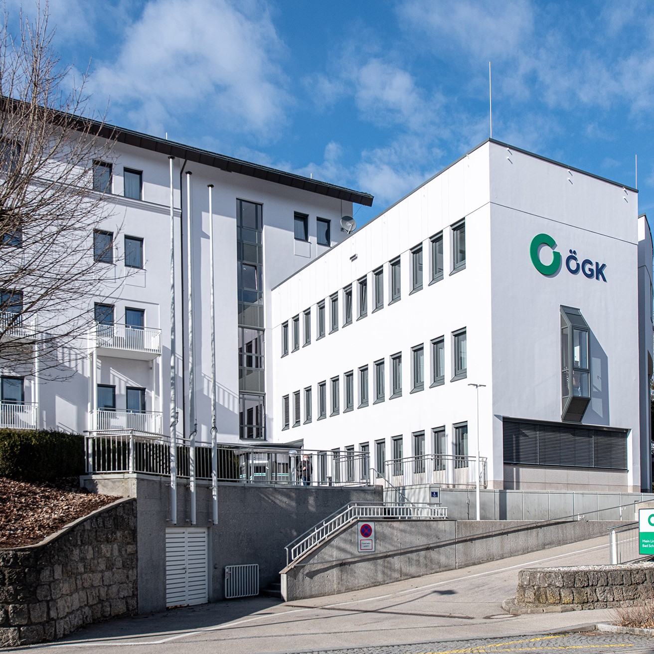 Au&szlig;enansicht des Gesundheitszentrums: Drei- beziehungsweise vierst&ouml;ckiger wei&szlig;er L-Bau; im Vordergrund eine Zufahrtstra&szlig;e und der Stiegenaufgang zum Haupteingang mit St&uuml;tzmauern und Handlauf;
