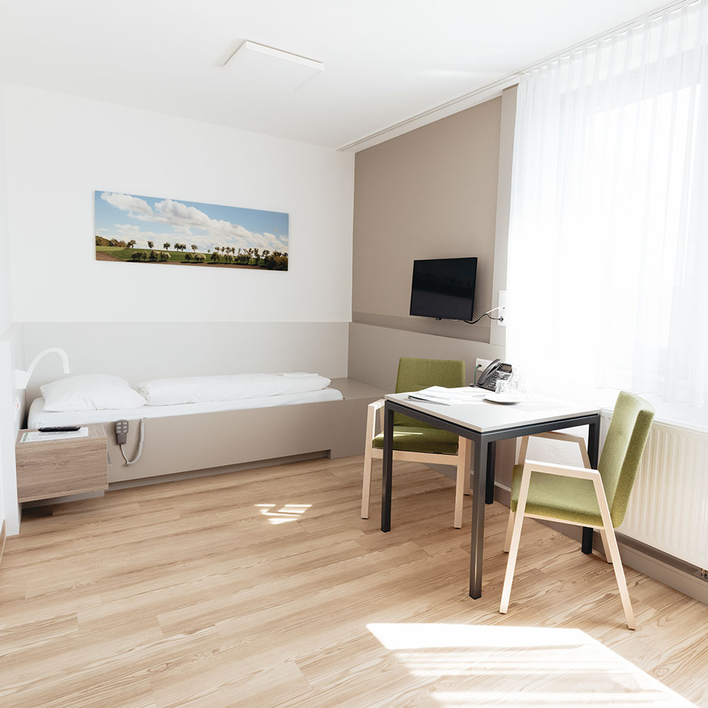 Helles Einbettzimmer mit Parkettboden; am Fu&szlig;ende des Bettes h&auml;ngt ein Fernseher an der Wand, neben dem Bett vor einem Fenster zwei gr&uuml;ne Sesseln und ein Tisch mit Telefon; gegen&uuml;berliegend neben dem Bett ein Nachtk&auml;stchen 