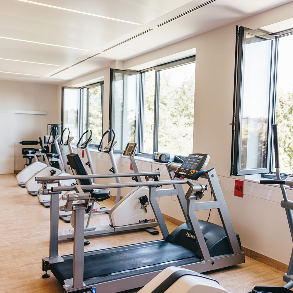 Fitnessraum im Gesundheitszentrum: Ein Laufband und verschiedene Ergometer stehen in einer Reihe vor einer breiten Fensterfront mit Blick ins Gr&uuml;ne. 