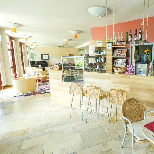 Ger&auml;umige Cafeteria mit Barhockern vor einem Tresen mit Snacks und Getr&auml;nken. Im Hintergund bequeme Polstersessel an kleinen Tischchen.