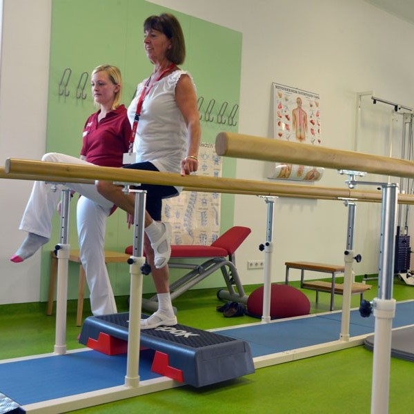 &Uuml;bung im Therapieraum: Eine Frau steht einbeinig im Parellelgehbarren auf einem Sockel, das andere Bein rechtwinkelig hochgehoben, die H&auml;nde ber&uuml;hren zur Sicherheit leicht die Holzbarren. Die Therapeutin macht die &Uuml;bung daneben mit.