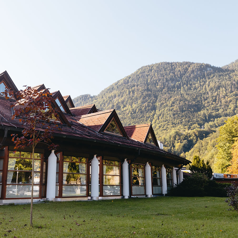 Au&szlig;enansicht des Speisesaalbereichs: vor der wintergartenartigen Fensterfront mit wei&szlig;en S&auml;ulen ist eine Wiese, ein Baum und Str&auml;ucher; Man sieht auch das Dach mit den Dreiecksgaupen und Blick in die gr&uuml;ne Berglandschaft.