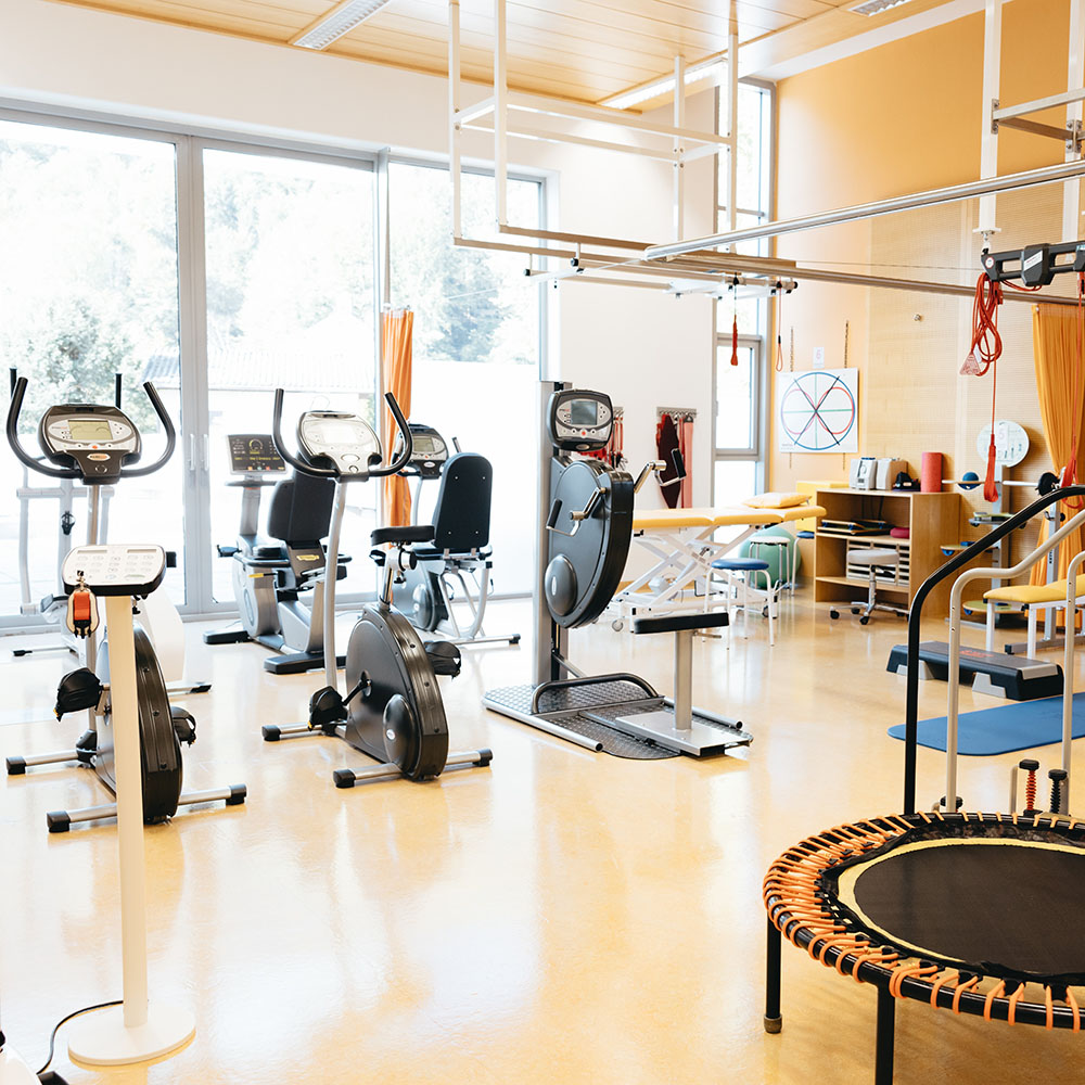 Gro&szlig;er Trainingstherapieraum im Gesundheitszentrum; breite hohe Fensterfront mit Blick ins Gr&uuml;ne, davor stehen in zwei Reihen jeweils drei Trainingsger&auml;te, rechts vorne ein Trampolin, rechts hinten im Bild ein mit Vorhang abtrennbarer Bereich mit Liege;