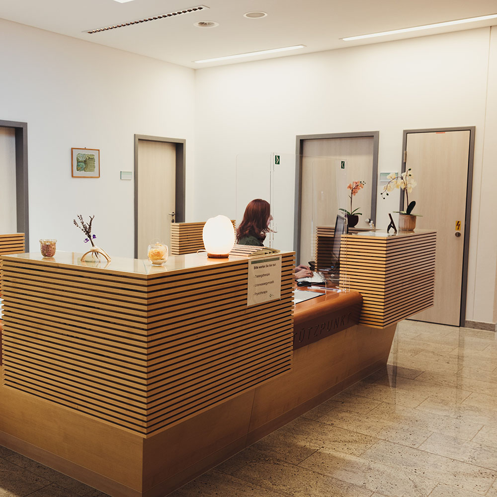 Anmeldebereich f&uuml;r Therapien: gro&szlig;e viereckige Rezeption mit Covid-Schutz-Glas vor dem Anmeldebereich; dahinter sitzt eine Frau und arbeitet am Computer; auf der Theke stehen Orchideen und andere Accsessoires; T&uuml;ren zu Therapier&auml;umen im Hintergrund;