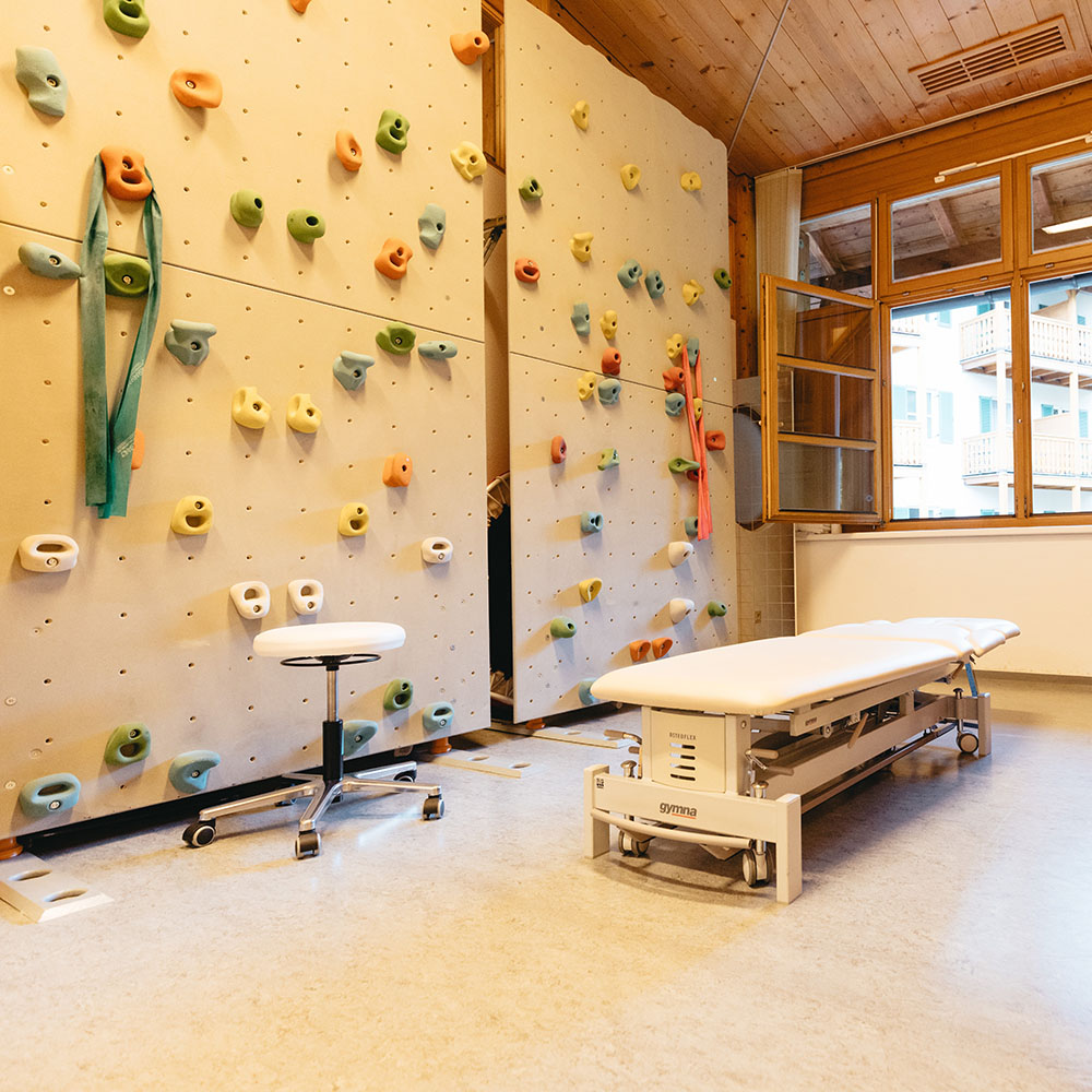 Gro&szlig;er Therapieraum, linkerhand mit therapeutischer Kletterwand, heller Boden, Holzdecke. Eine wei&szlig;e Behandlungsliege mit Hocker steht vor einem ge&ouml;ffneten Fenster mit Blick auf eine Geb&auml;udefront.