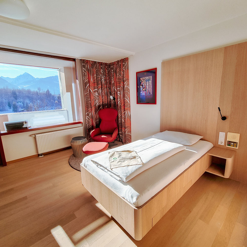 Helles Zimmer mit einem Bett, einem Sessel, Holzfu&szlig;boden und einem fenster mit Blick auf Wald und Berge.